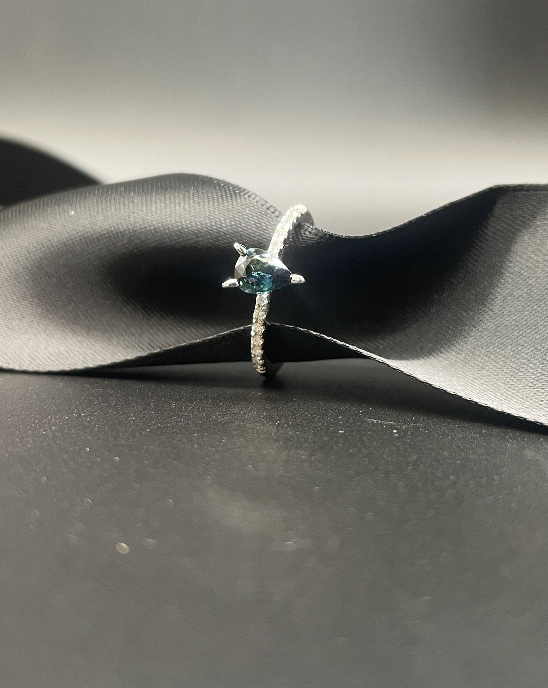 The Calista engagement ring in white gold with a pear shaped teal sapphire and black ribbon with a dark background.