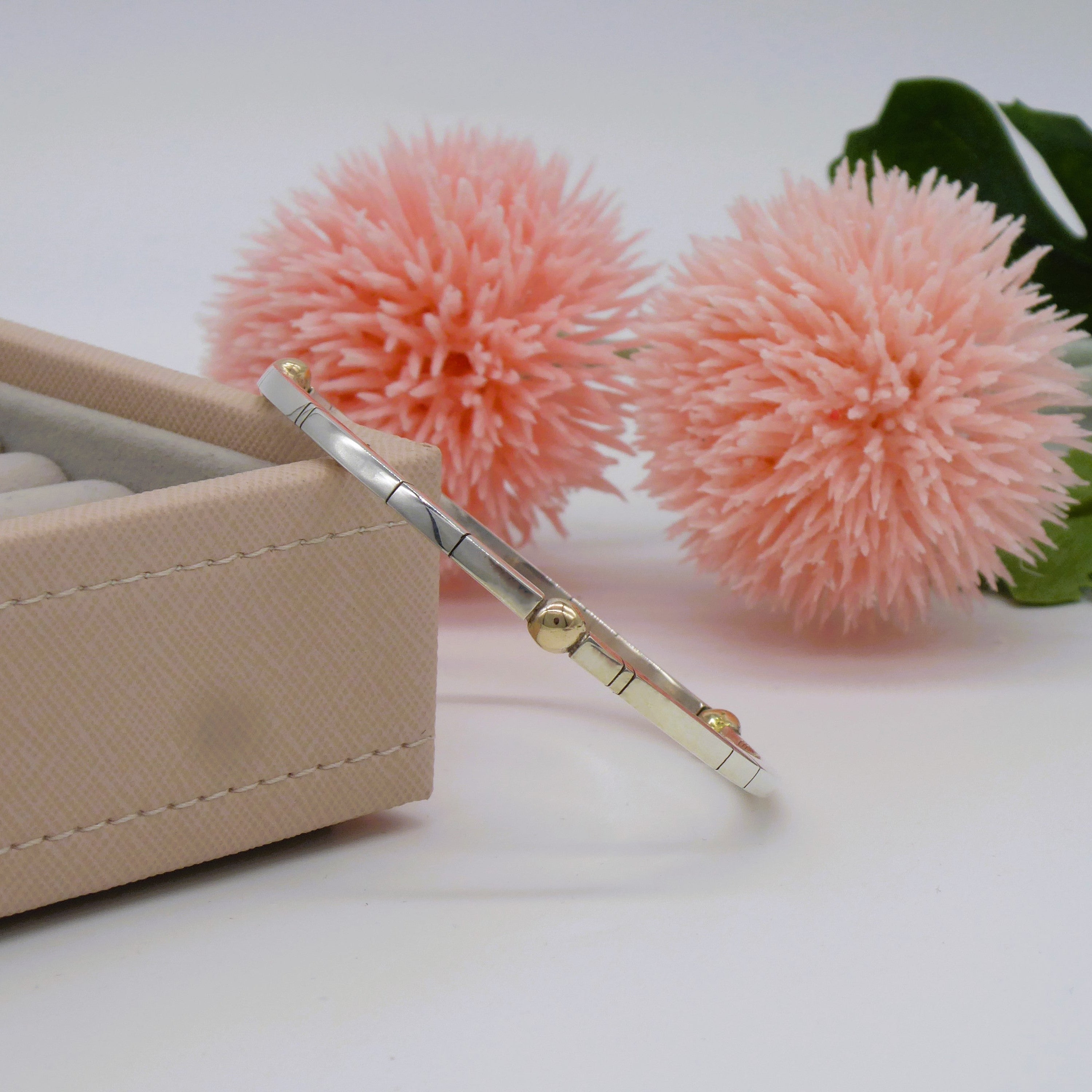 A silver and gold bracelet on a white surface with pink flowers and a jewellery box in the background.