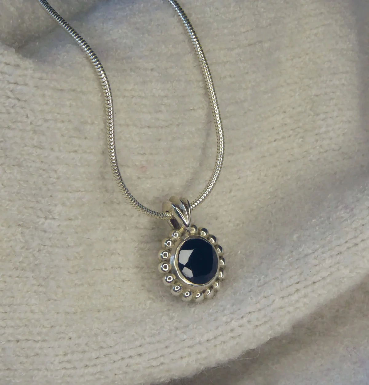 Angled close-up of Alto Majestic black spinel and recycled silver pendant on a cream knit fabric, showing the brilliant-cut black stone and millgrain beaded silver frame