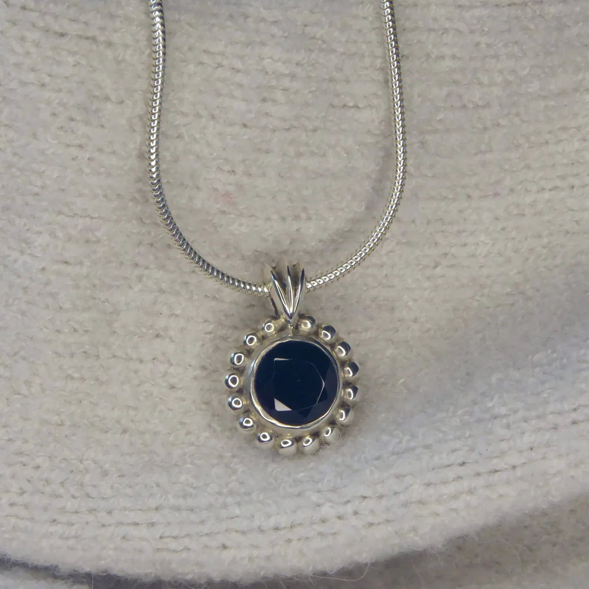 Alto Majestic black spinel and recycled silver pendant resting on a cream knit fabric, front view showing deep inky stone and millgrain beaded border on a snake chain