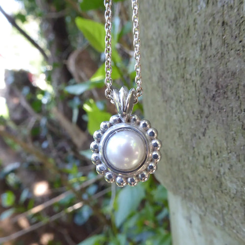 White freshwater pearl and recycled silver Alto Pearl pendant hanging against a stone wall with garden foliage, showing soft pearl lustre and millgrain beaded silver setting on a trace chain