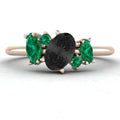 Front view of the Atlantis engagement ring, featuring a central pear-shaped black diamond surrounded by an asymmetrical cluster of vivid green emeralds and white diamonds in 18ct rose gold.