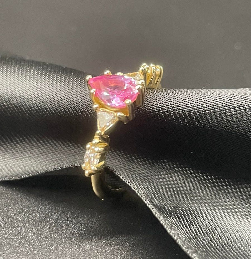 A bespoke version of the Maisie engagement ring set with a pear shaped pink sapphire, diamonds, yellow gold with black ribbon and a dark background.