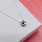 Silver necklace with a blue sapphire gemstone pendant on a white surface with a pink background