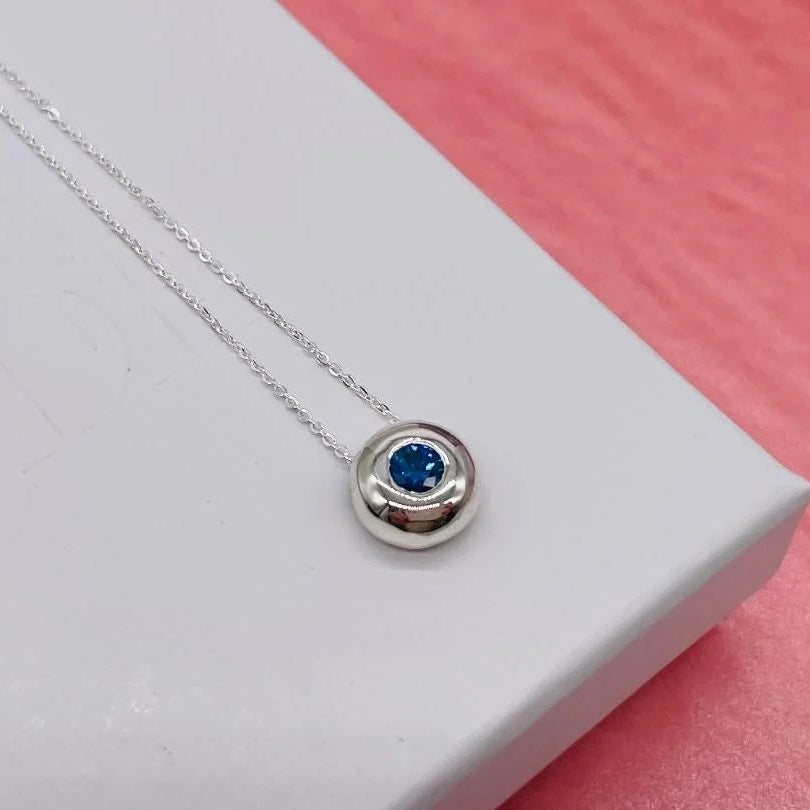 Silver necklace with a blue sapphire gemstone on a white box with a pink background
