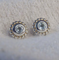 A pair of sterling silver stud earrings featuring round pale sky blue topaz gemstones surrounded by a textured millgrain border.