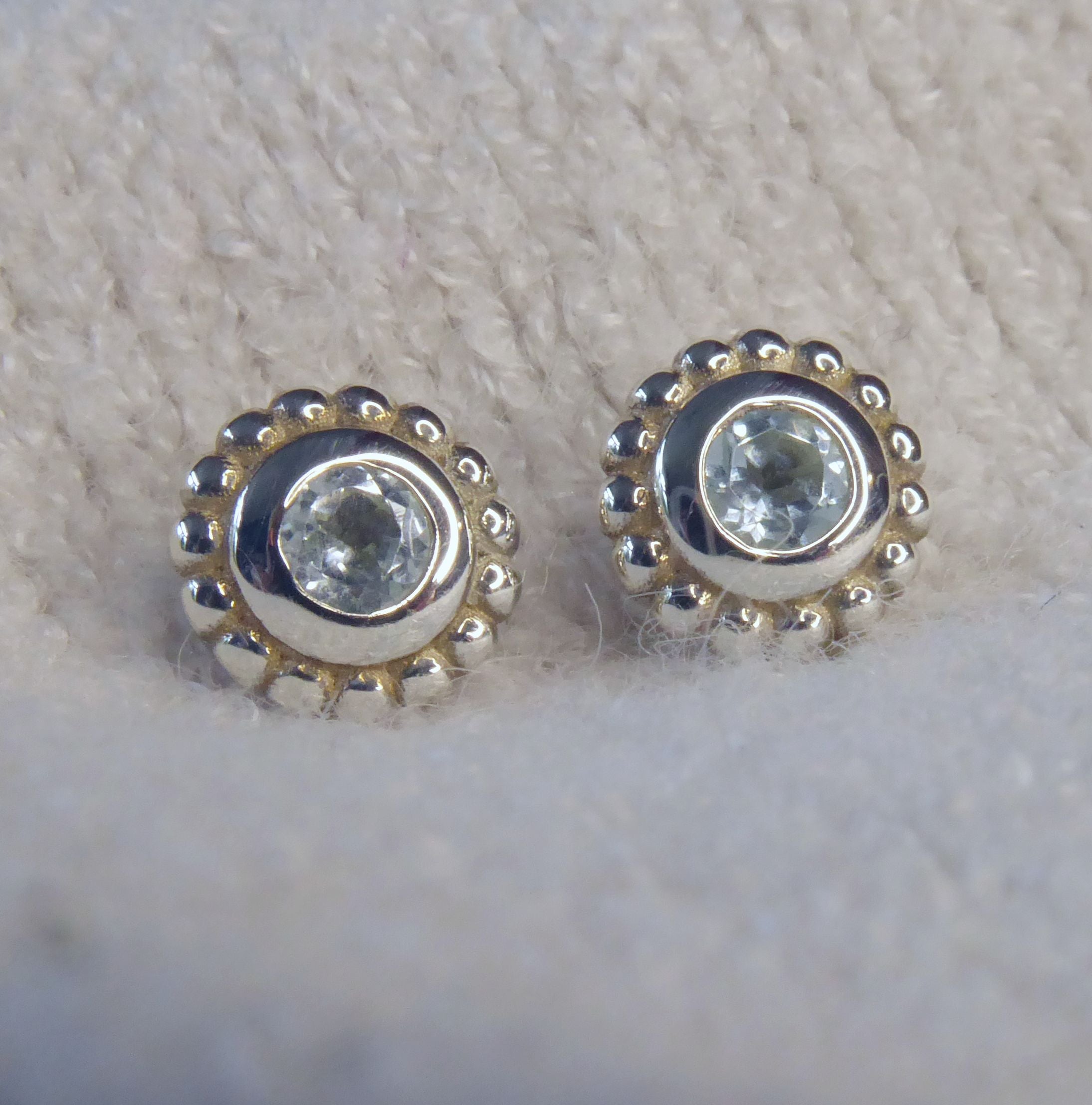 A pair of sterling silver stud earrings featuring round pale sky blue topaz gemstones surrounded by a textured millgrain border.