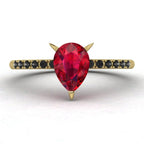 Top-down studio hero shot of the Calista engagement ring, featuring a vibrant pear-shaped red ruby on an 18ct yellow gold band with black diamond shoulders.