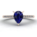 Front studio hero shot of the Calista engagement ring, featuring a prominent pear-shaped blue sapphire on a rose gold band with sparkling white diamond shoulders.