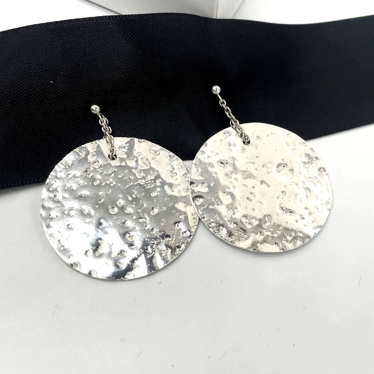 Pair of medium Glitter Ball recycled sterling silver disc earrings resting on a black satin ribbon, showing hand-textured shimmer surface and chain drops