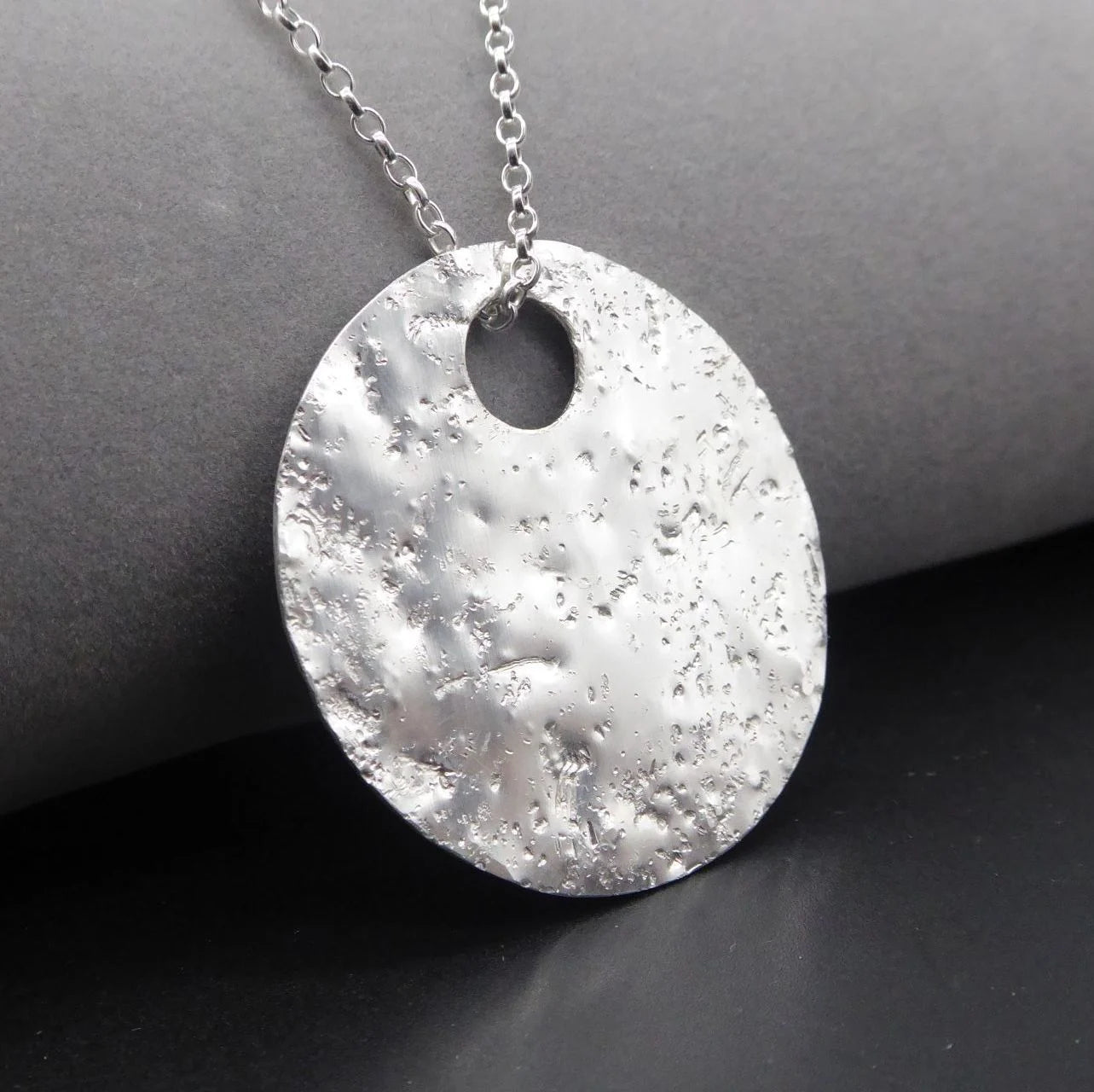 Silver handmade textured pendant necklace on a dark surface - slight side angle