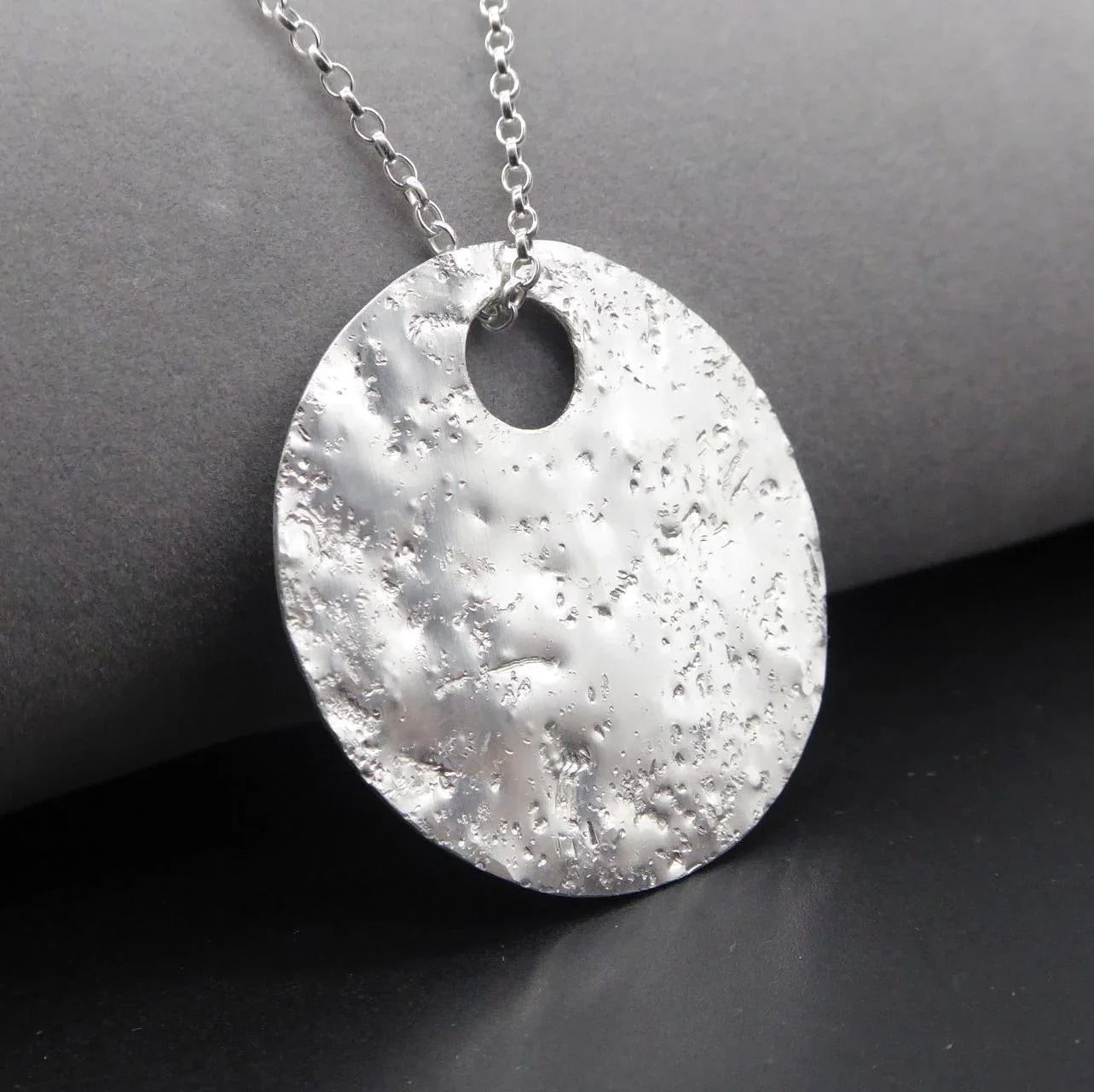 Angled view of hand-textured recycled sterling silver Glitter Ball disc necklace on a dark grey surface, showing the pendant's depth and circular eyelet detail