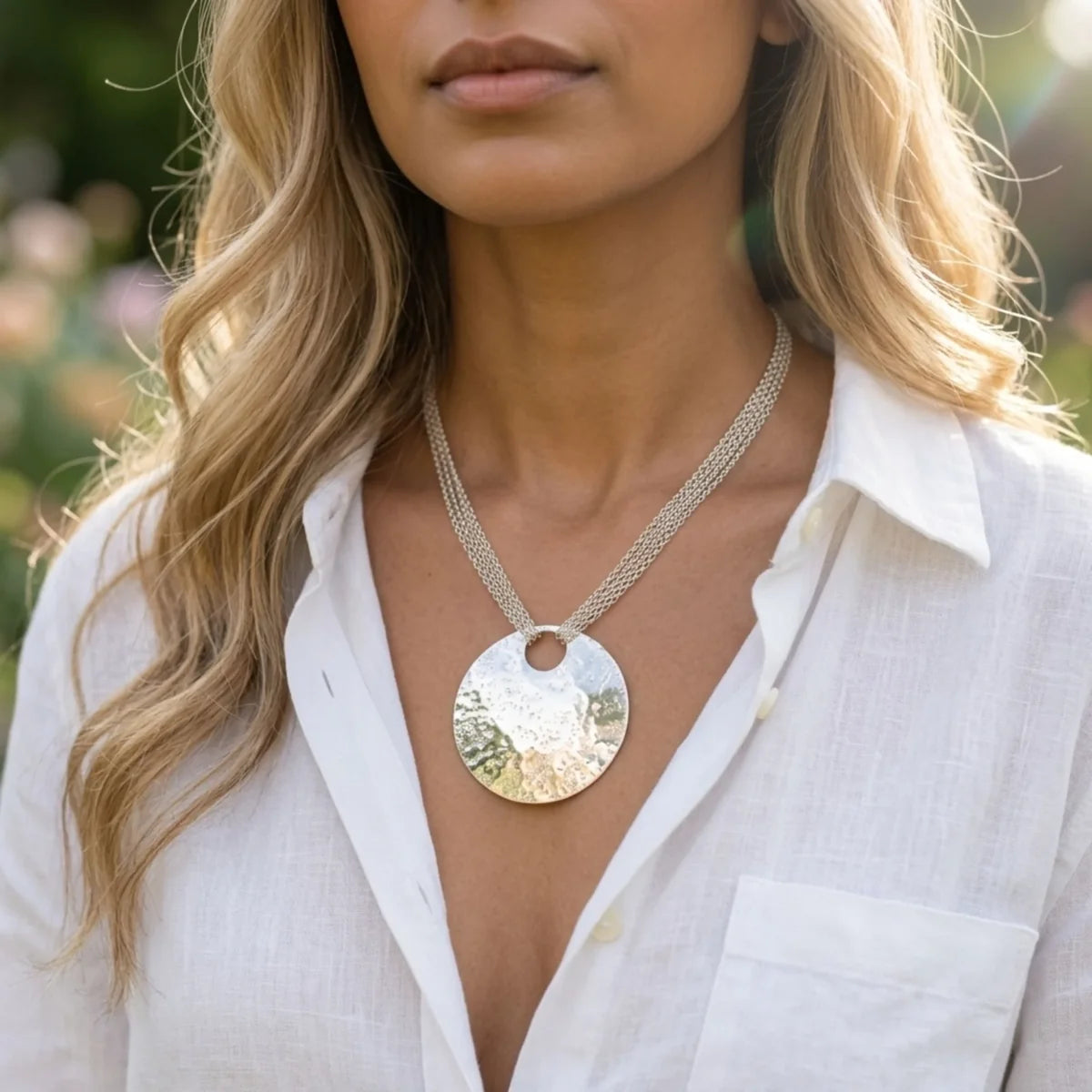 Model wearing the Glitter Ball Multi Chain recycled silver statement necklace outdoors, showing large hand-textured disc pendant on multi-strand chain with a white linen shirt