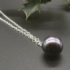 Close-up of a dark grey-black freshwater pearl pendant on a sterling silver chain, resting on a charcoal surface beside green leaves.
