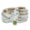 A multi-layered wave-style silver ring featuring textured silver bands, polished gold-toned accents, and a small round green sapphire gemstone, shown on a plain white background.