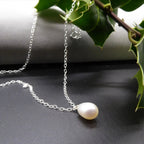 White pear-shaped freshwater pearl pendant and silver chain arranged on a dark surface beneath holly leaves.
