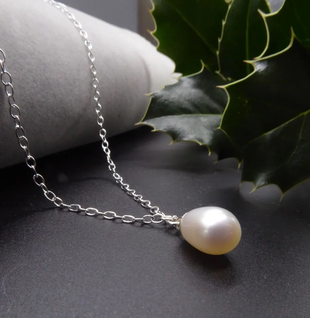 White pearl drop necklace with a silver chain draped over a grey display beside green holly leaves.