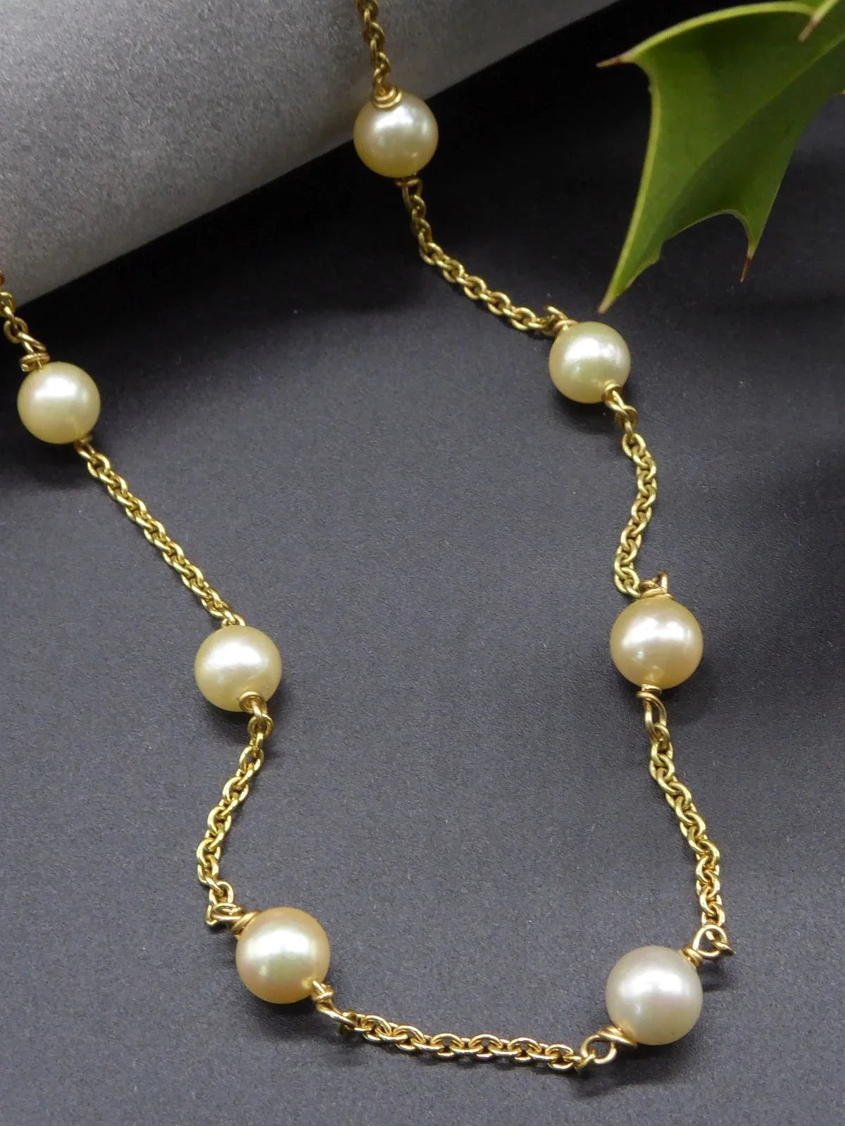Ivory freshwater pearls spaced along an 18ct yellow gold chain necklace, arranged on a dark grey surface beside a green leaf.