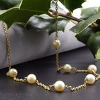 Seven ivory freshwater pearls spaced along an 18ct yellow gold chain, draped over a grey display with holly leaves beside it.