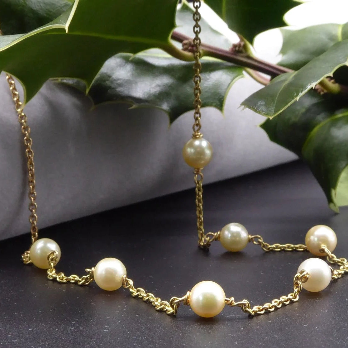 Seven ivory freshwater pearls spaced along an 18ct yellow gold chain, draped over a grey display with holly leaves beside it.