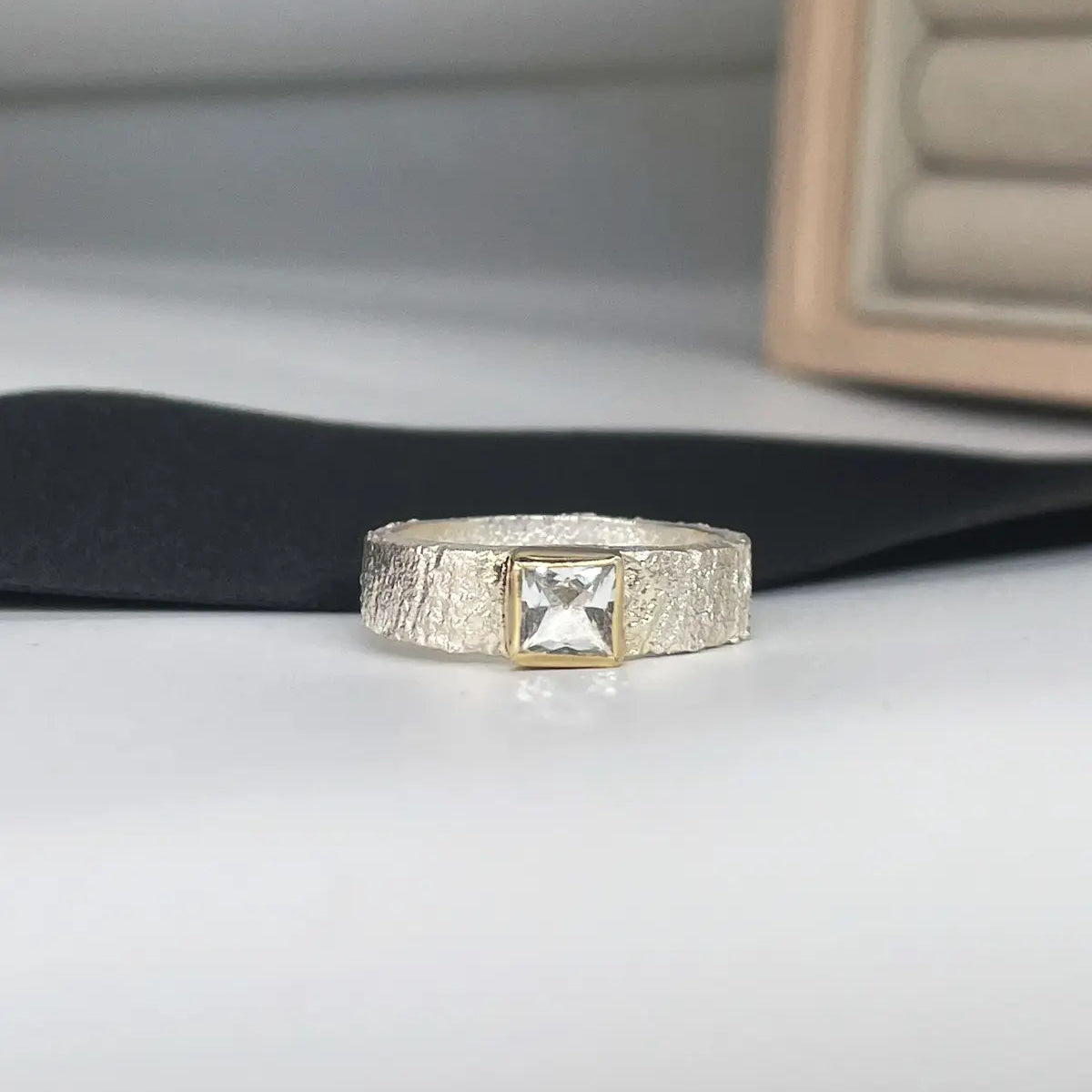Rivda ring with a square princess-cut white topaz in a yellow gold setting, showing its wide, organically textured silver band on a pale surface.