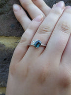 Selene oval teal sapphire and diamond asymmetric cluster engagement ring worn on hand in 18ct white gold, showing the deep blue-green sapphire and diamond cluster, handmade in our London studio