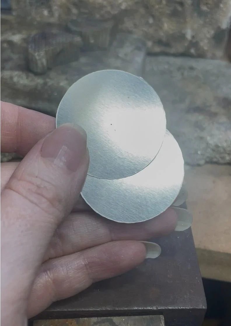 Dividers scribing circles onto a blue-coated recycled silver sheet in the London jewellery studio, marking out earring discs ready for cutting