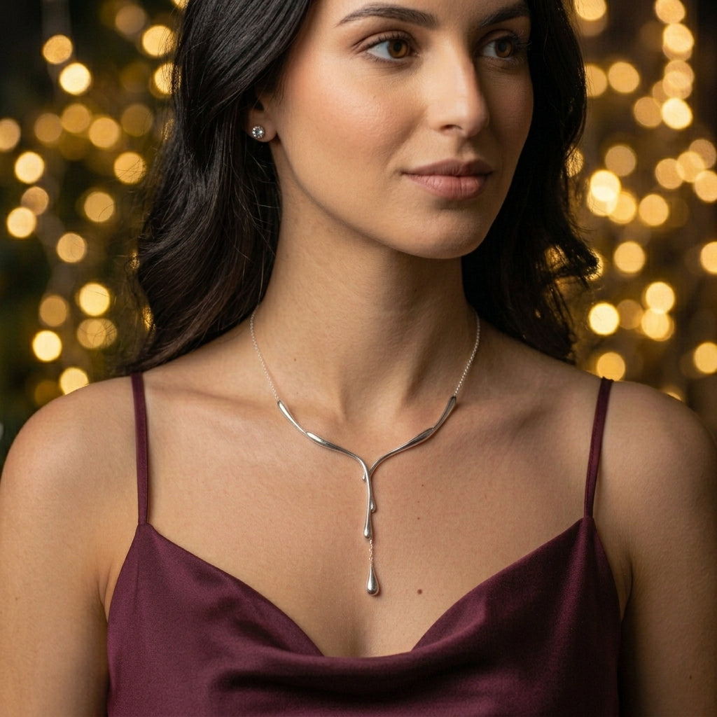Model wearing a silver drip necklace with a dangling teardrop pendant and a burgundy satin top, against softly blurred golden festive lights.