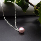 Pink freshwater pearl pendant on a sterling silver trace chain, displayed on a dark surface beside green leaves.
