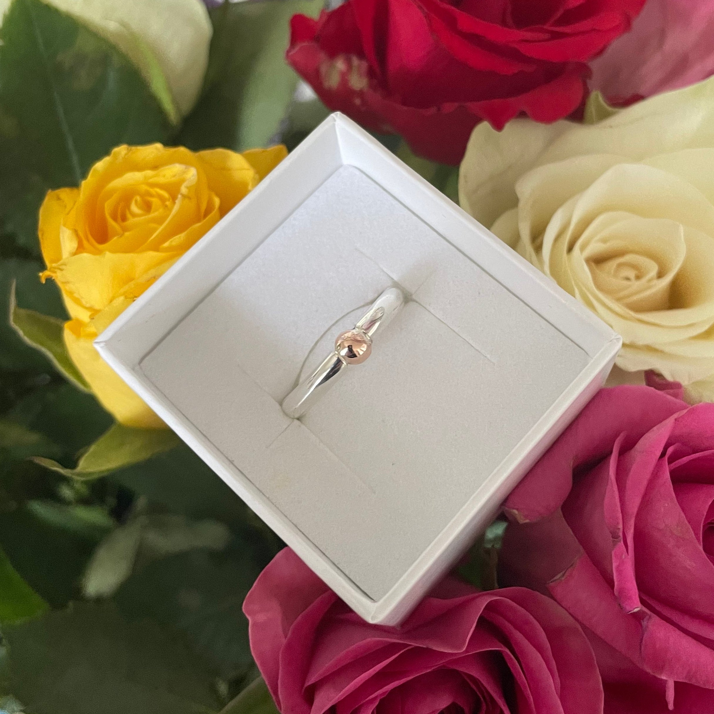 Silver and gold ring in a box surrounded by colourful roses with a blurred background