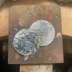 Two hand-textured recycled silver discs resting on a steel bench block in the London studio, showing the glitter ball surface texture before finishing