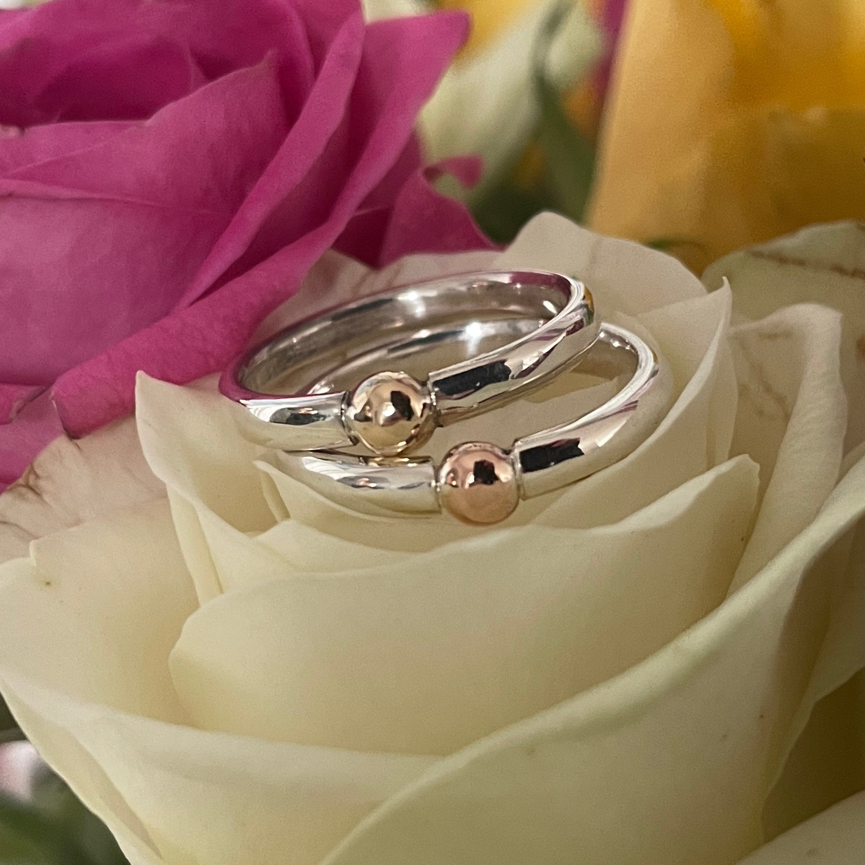 Two silver rings with gold accents on a white rose with pink flower in the background
