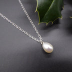 Angled close-up of a lustrous white pearl drop pendant on a sterling silver chain beside a grey display and holly leaves.